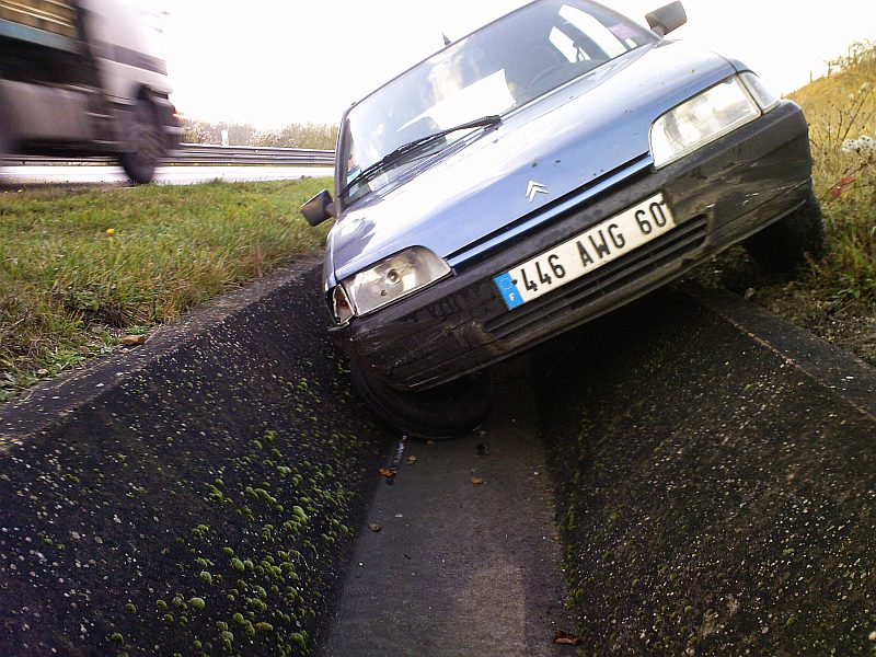 La Citroën AX bleue, capot en avant, immobilisée dans la rigole en béton qui borde la chaussée ; un poids lourd passe à gauche.