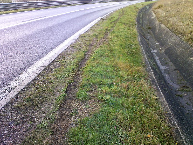 La chaussée et sa rigole vues vers l’horizon ; des traces de pneus marquent l’herbe du bas-côté sur plusieurs mètres.
