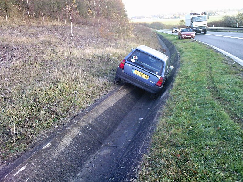 Vue de l’arrière : la voiture couchée dans la rigole ; plus haut, un poids lourd et une voiture circulent normalement sur la route.