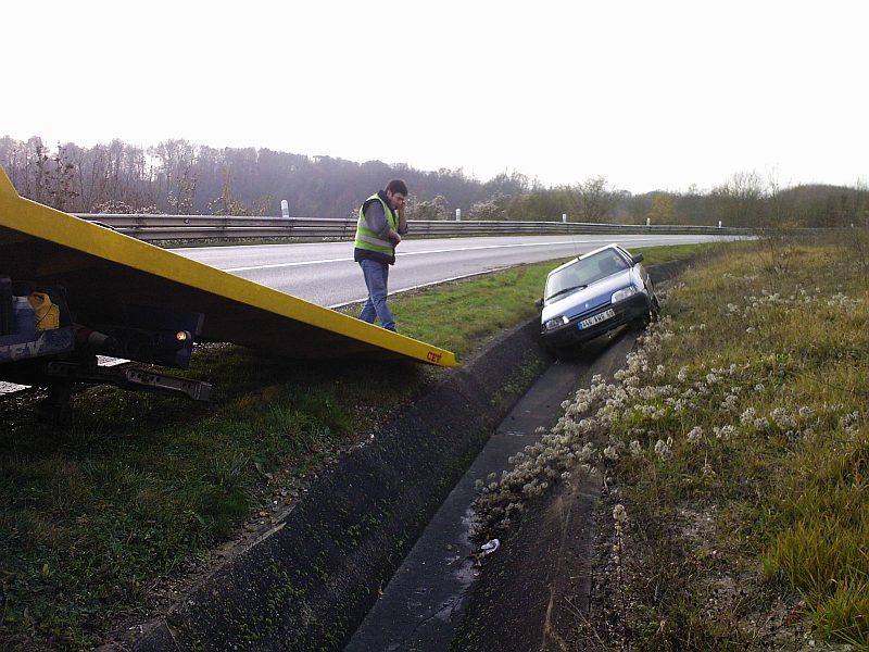 La rampe jaune du camion de dépannage barre le premier plan ; un homme en gilet de haute visibilité patiente au bord de la chaussée, la voiture toujours dans la rigole.